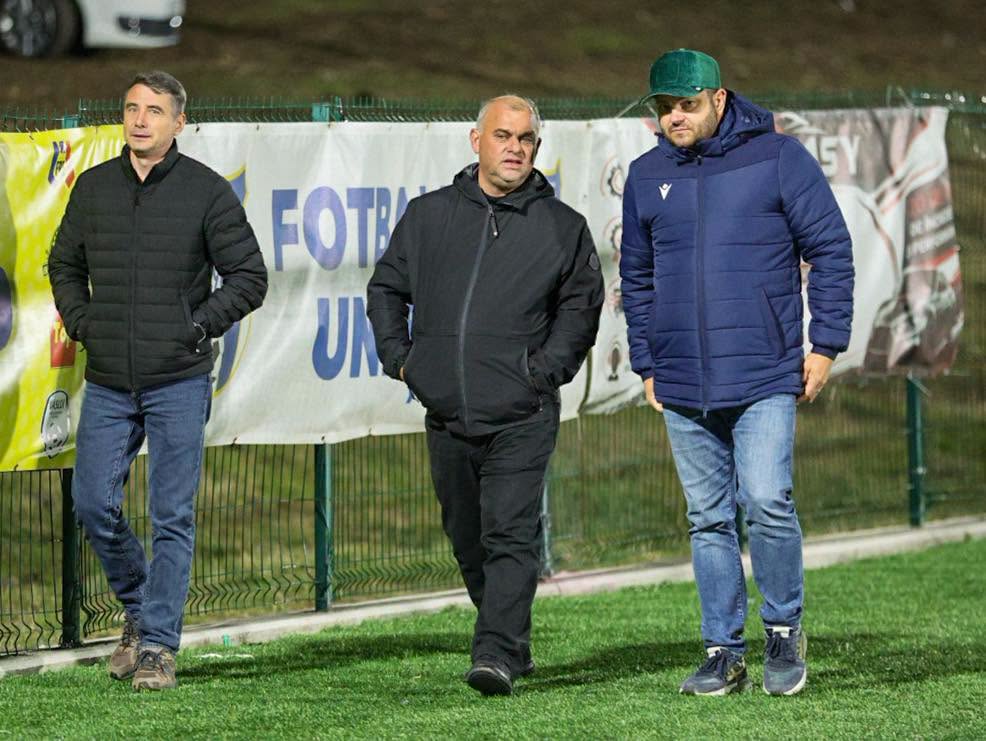 Trei bărbați îmbrăcați în jachete de toamnă, surprinși mergând pe marginea unui teren de fotbal din Huși, în timpul Cupei Hușana 2026.